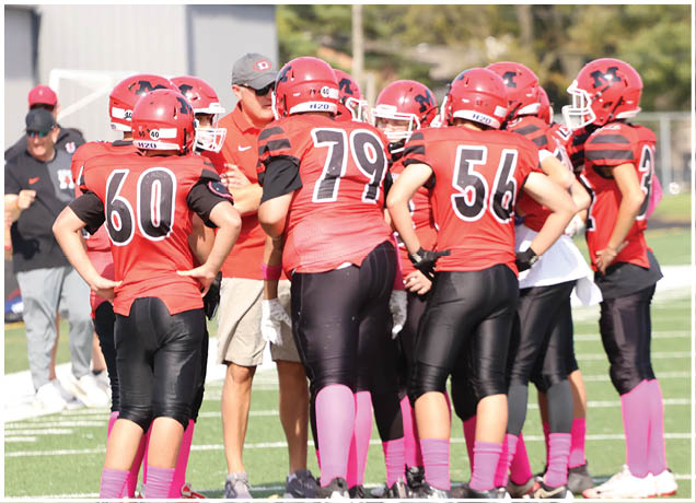 A group of people wearing red and black football uniforms with pink socks are gathered on a grassy field, seemingly listening to a coach in casual clothing. Description generated by AI