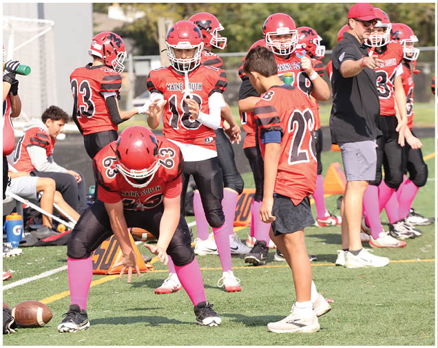 A group of people wearing red football uniforms with pink socks are gathered on a field, with one person in a crouched position preparing for a play and another person standing nearby. Description generated by AI