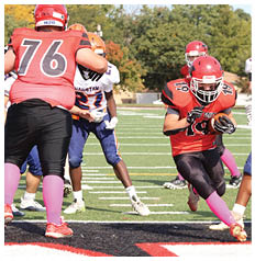 A person in an orange and black football uniform with pink socks is running with the ball while other players in blue and orange uniforms attempt to block or tackle on a football field. Description generated by AI