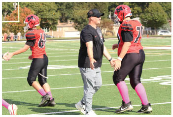 Two people stand on a football field, one wearing gray pants and a black shirt and the other wearing black athletic pants, pink socks, and a red jersey. Description generated by AI