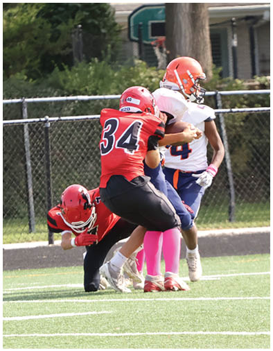 Several people wearing football helmets and uniforms are engaged in a tackle on a grassy field near a chain-link fence with houses in the background. Description generated by AI