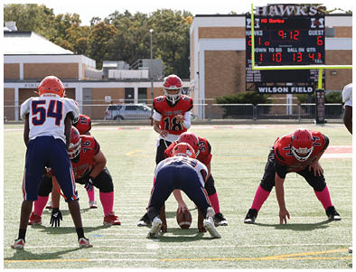 People in football uniforms are positioned on a field with a scoreboard in the background showing the home team leading. Description generated by AI