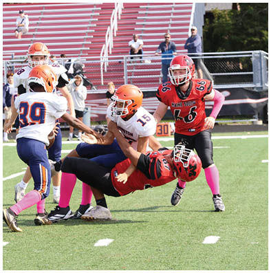 Children playing American football on a field with some players tackling and others standing nearby wearing colorful uniforms and pink socks. Description generated by AI