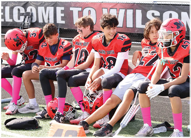 A group of young people in red and black football uniforms with pink socks are sitting on a bench on a sports field, some holding helmets and one using crutches. Description generated by AI