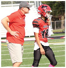 A person wearing a red football jersey with the number 18, black pants, and pink socks is standing on a green football field. Description generated by AI