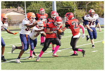 Several people in football uniforms are actively engaged in a game on a grassy field, with one person in a red uniform holding the ball while others in white and blue uniforms attempt to tackle or block. Description generated by AI