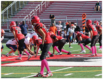 A group of people wearing red and black football uniforms with pink socks are lined up on a football field facing an opposing team in white uniforms, preparing for a play. Description generated by AI