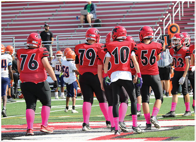 People wearing red football uniforms with pink socks are standing on a football field near the sideline with others in white and orange uniforms in the background. Description generated by AI