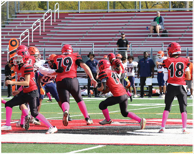Children wearing red helmets and jerseys with pink socks are playing football on a field with empty bleachers and a few adults in the background. Description generated by AI