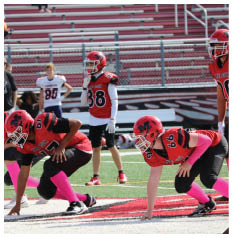 A person in a red football uniform and helmet stands on a field with other players and people in the background near empty bleachers. Description generated by AI