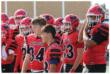 A group of people wearing red and black football uniforms stand on a field near a person in a red vest labeled student who is gesturing with one hand. Description generated by AI