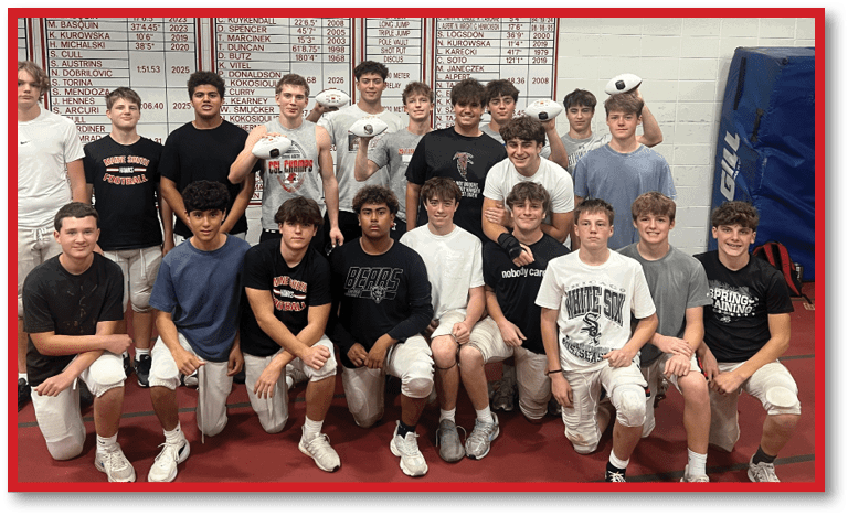 A group of people wearing athletic clothing and knee pads pose together indoors in front of a scoreboard and gym equipment. Description generated by AI