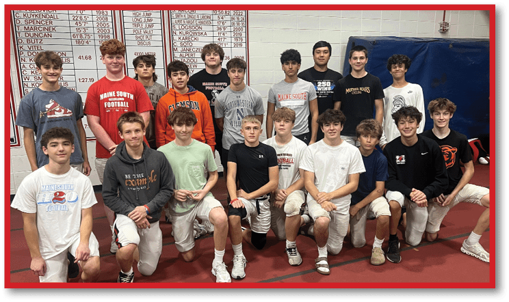 A group of people wearing casual sportswear and shorts pose together indoors on a red floor with a scoreboard visible in the background. Description generated by AI
