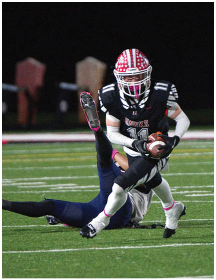 A person wearing a red and white helmet and black sports uniform is running with a football while another person in a dark uniform is falling on the turf field near a goalpost. Description generated by AI