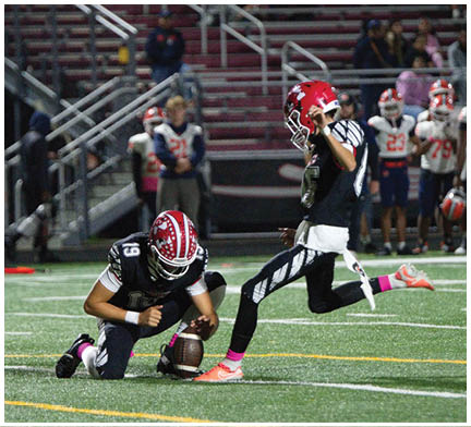 Two people in football uniforms are on a field with one holding the ball on the ground while the other is kicking it, with spectators and other players in the background. Description generated by AI