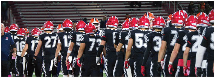 People wearing black sports uniforms with red helmets and pink socks stand in a line on a red and green field. Description generated by AI