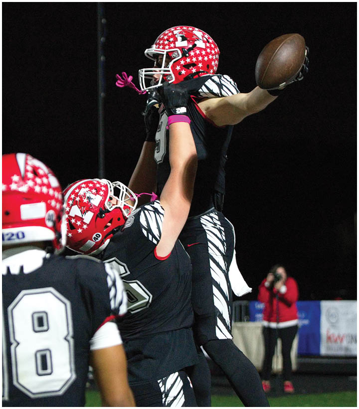 Three people wearing black and white football uniforms with red helmets celebrate on a field at night, with one person lifting another who holds a football. Description generated by AI