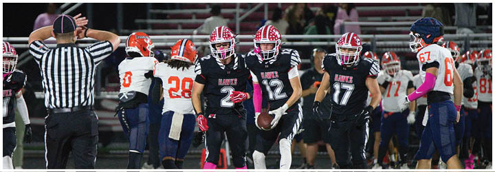 People in football uniforms are on a field with a referee signaling in the background during a game. Description generated by AI