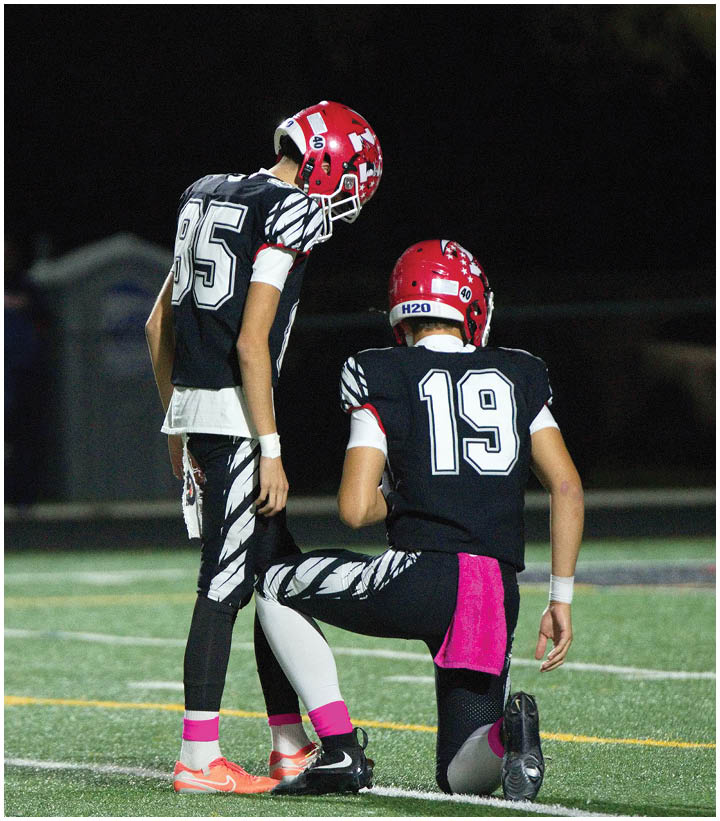 Two people wearing black and white football uniforms with red helmets are on a field at night, with one standing and the other kneeling. Description generated by AI