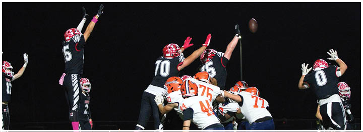 Several people in football uniforms are actively jumping and reaching to catch or block a football during a nighttime game. Description generated by AI