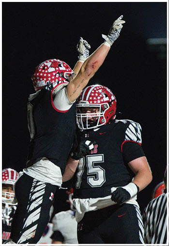 Two people wearing black football uniforms with red and white star-patterned helmets celebrate on a field at night. Description generated by AI