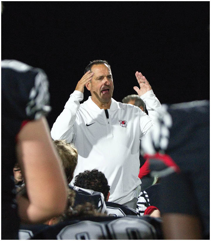 A person in a white jacket is speaking animatedly to a group of people wearing black and white sports uniforms with numbers on the back, likely during a nighttime sports event. Description generated by AI