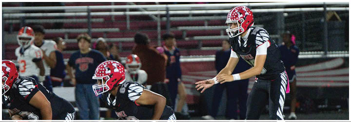 People in football uniforms with pink socks are positioned on a field, preparing for a play during a game. Description generated by AI