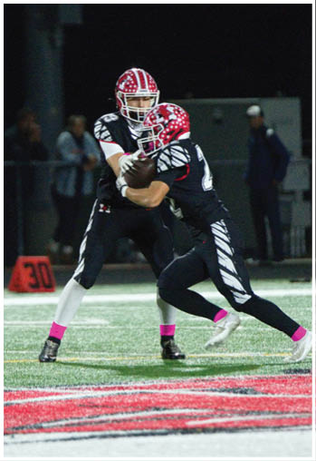 Two people wearing black football uniforms with red helmets and pink socks are engaged in a play on a football field at night. Description generated by AI