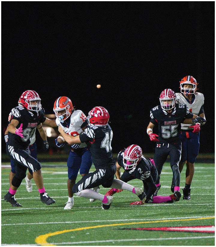 A group of people wearing football uniforms are actively playing on a field at night with one person holding the ball while others attempt to tackle or block. Description generated by AI