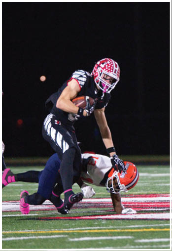 A person in a black and red football uniform is running with the ball while another person in an orange helmet and white jersey is on the ground attempting to tackle them on a football field at night. Description generated by AI