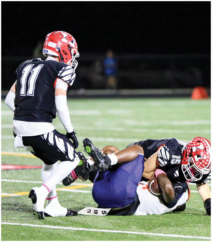 Two football players in black uniforms with red helmets tackle a player in a white uniform holding a football on a grassy field. Description generated by AI
