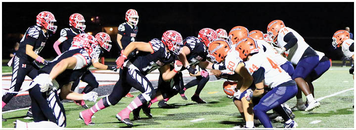 Two opposing American football teams in helmets and uniforms are lined up facing each other on a field at night, preparing for a play. Description generated by AI