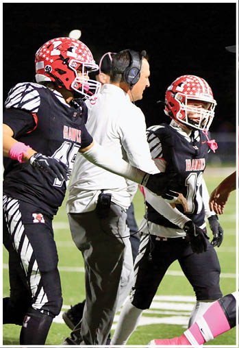 A group of people on a football field includes two players in black uniforms with red helmets and a person in a white shirt and headset interacting with them. Description generated by AI