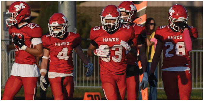 Four people in red football uniforms with helmets walk together on a field near an orange yard marker. Description generated by AI