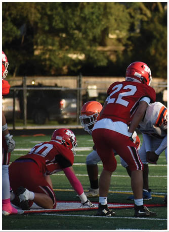A person wearing a red sports jersey with the number 28 and an orange helmet is outdoors near a fence with trees and a vehicle in the background. Description generated by AI