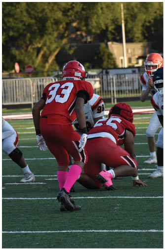 Two people in red football uniforms with pink socks are positioned on a football field facing opponents in white and orange uniforms during a game. Description generated by AI