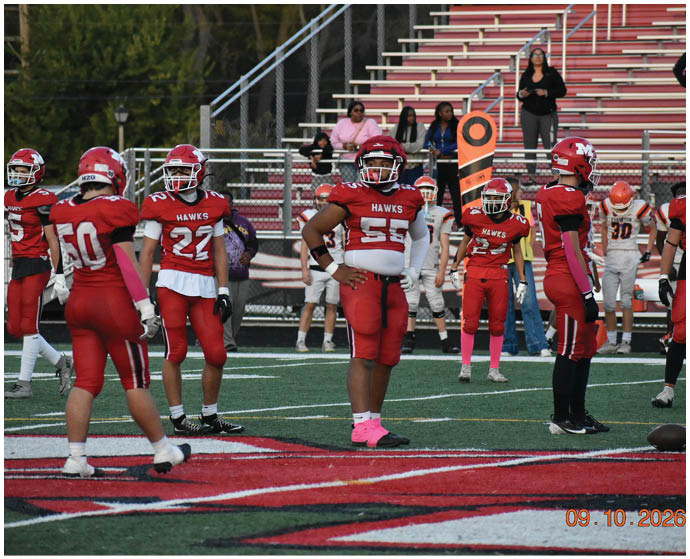 Several people in red football uniforms stand on a field near a large red and white logo with spectators in the background. Description generated by AI
