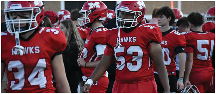 People wearing red football uniforms with the word “HAWKS" and numbers on the jerseys are standing together, likely preparing for a game. Description generated by AI