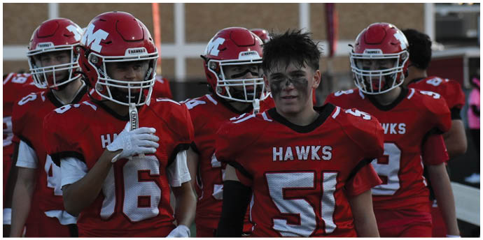 A group of people wearing red football helmets and uniforms stand together on a field with a building in the background. Description generated by AI