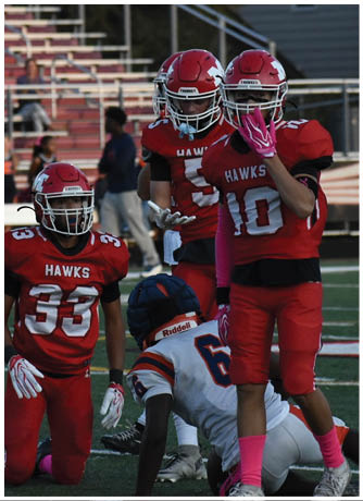 Three people wearing red football uniforms and helmets with the word “HAWKS" on their jerseys are standing on a field with bleachers in the background. Description generated by AI