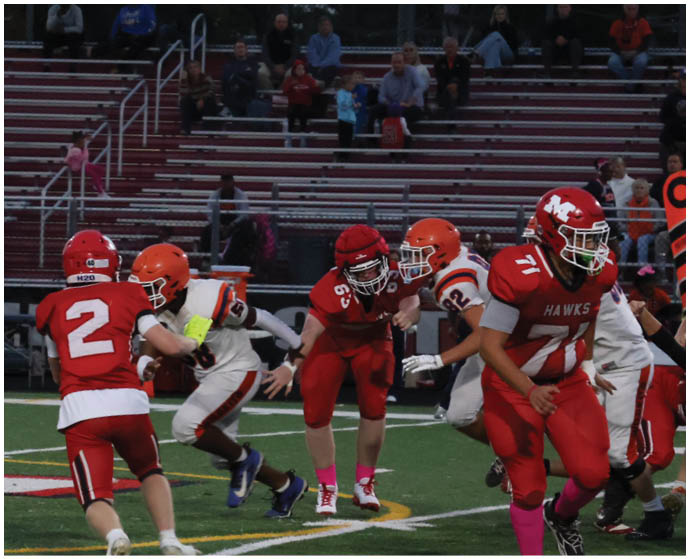 People in red and orange football uniforms are playing on a field with bleachers and spectators in the background during a game. Description generated by AI