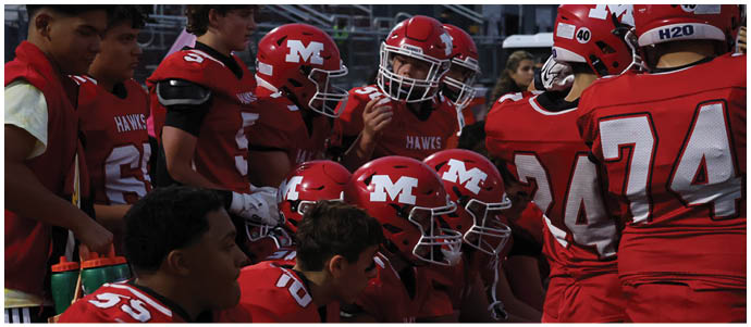 Several people wearing red football uniforms and helmets are sitting closely together, appearing focused and attentive. Description generated by AI