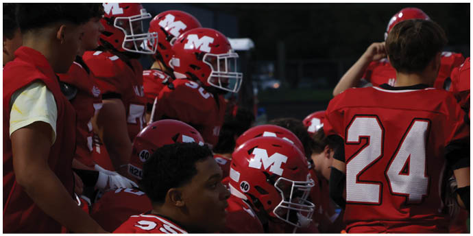 A group of people wearing red football uniforms and helmets are gathered closely together, with one person standing and others sitting or crouching. Description generated by AI