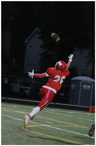 A person in a red football uniform leaps with arms outstretched attempting to catch a football while another person in a white football uniform runs nearby on a field at dusk. Description generated by AI