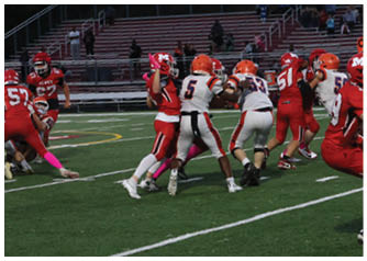 Players in red and white football uniforms are actively engaged in a game on a green field with empty bleachers in the background. Description generated by AI
