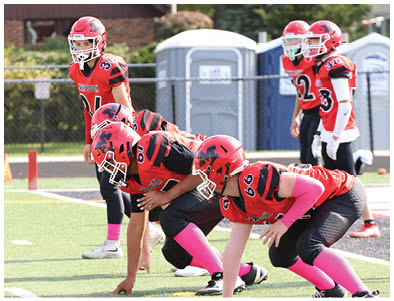 Several people wearing red football uniforms and helmets are positioned on a field preparing for a play, with some crouched in a stance and others standing behind them. Description generated by AI