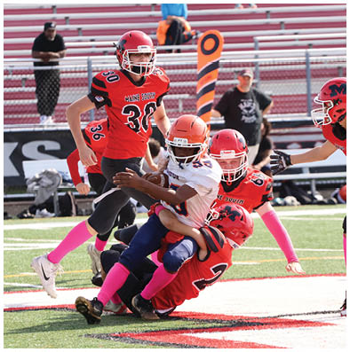 Four people in football uniforms with pink socks are tackling a person in a white jersey and orange helmet on a football field. Description generated by AI