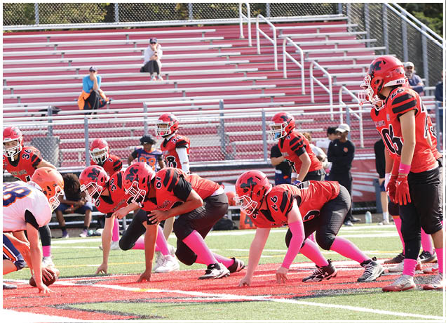 Two youth football teams in orange and red uniforms face each other on the field in a ready stance with empty bleachers in the background. Description generated by AI