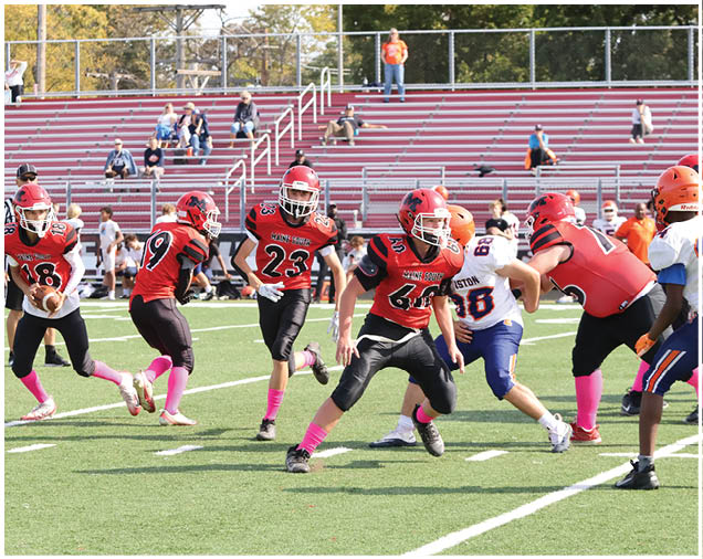 Adults in red and black football uniforms with pink socks are playing on a green field while adults in white and blue uniforms attempt to block them. Description generated by AI