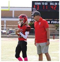A person wearing a red polo shirt and light-colored shorts stands on a sports field near another person in a sports uniform. Description generated by AI
